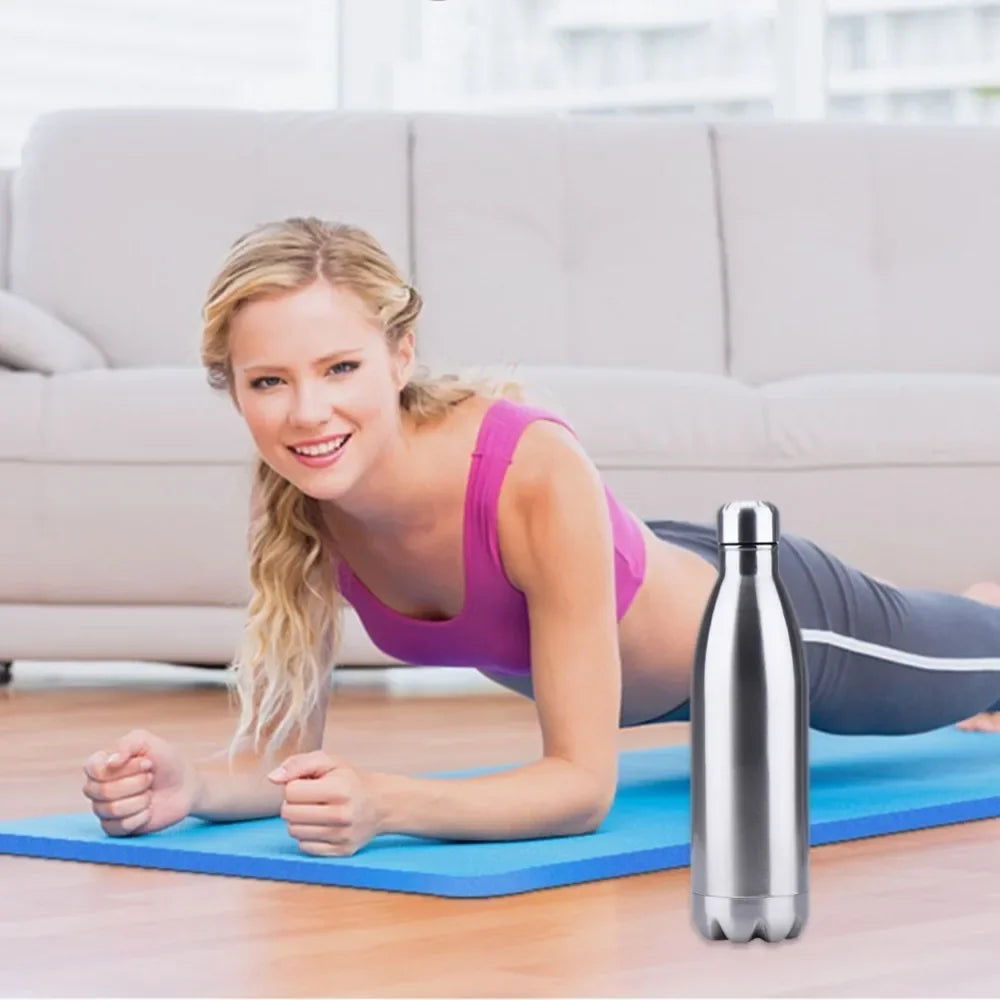 Une femme en tenue de sport sourit en faisant une planche sur un tapis de yoga bleu à l'intérieur, avec la Gourde en métal | Fit™ de Mon Univers Sport, connu pour son étanchéité avancée, placé au premier plan.