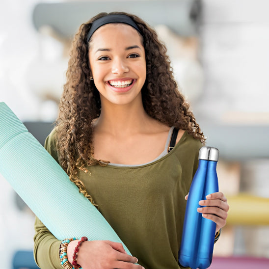 Une jeune femme aux cheveux bouclés sourit, tenant la gourde en métal Fit™ de Mon Univers Sport. Vêtue d'un haut vert et de bracelets à perles, elle admire sa conception étanche et sa durabilité au milieu des équipements d'exercice en salle.