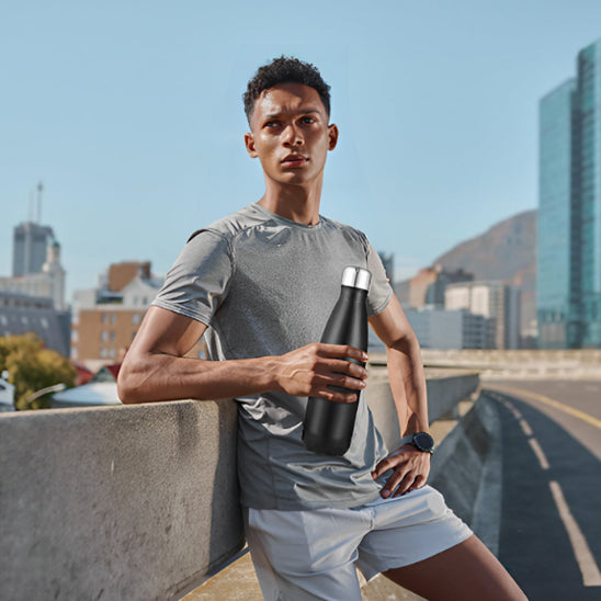 Un homme vêtu d'une chemise de sport grise et d'un short blanc se tient sur un toit, tenant la Gourde en métal | Fit™ de Mon Univers Sport, avec des bâtiments de la ville et une montagne en arrière-plan.